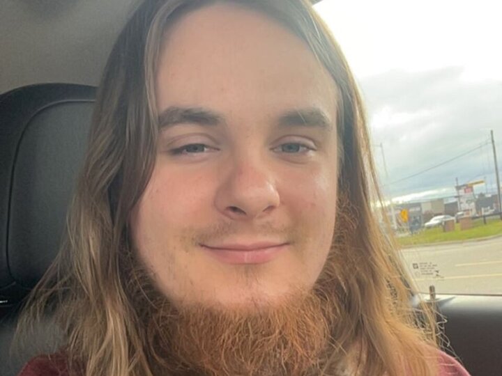 Young man with long hair and a beard smiling inside a car, cloudy sky visible through the window.