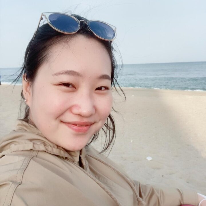 Youmin Kim - smiling, sunny but cooler day at the beach as she is wearing a light tan parka, sunglasses lifted sitting on the top of her forehead;; we see looking at us turned to her right shoulder, body facing to the side, smiling pleasantly