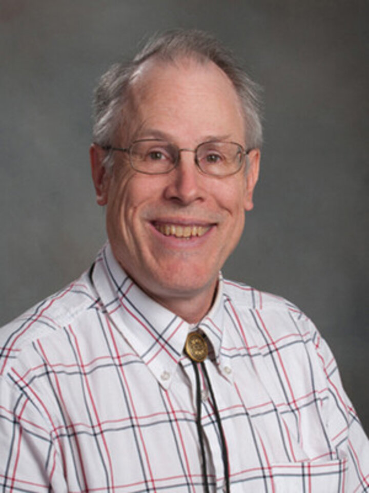 Charles (Chuck) Riedesel - stock department photo - he's wearing a light shirt with light red, blue very thin lined squaring pattern - and a bolo tie - smiling