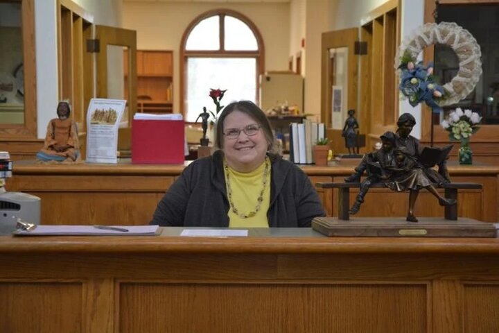 Laureen Riedesel centered in the wide shot photo -- yellow shirt with dark cardigan, slight smile, -- she is seated behind the front desk of the Beatrice Public Library where she retired as director - lots of library like things around -- space is large with a nice arched window far behind her letting in the blue sky and light of the day